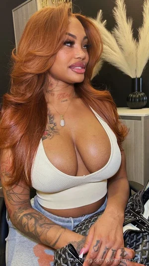 Woman with long red hair, tattoos, wearing white crop top and jeans, sitting indoors.