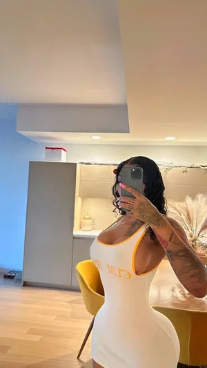 Woman in white tank top with yellow trim, tattoos on arms, taking mirror selfie in modern kitchen.