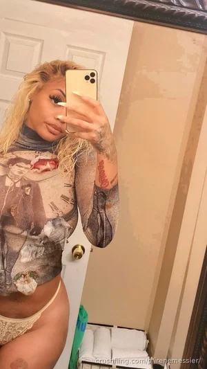 Woman with blonde hair and tattoos wears sheer top and beige underwear, taking mirror selfie in
