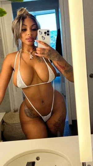 Woman in white bikini taking mirror selfie, tattoos visible, indoor setting.