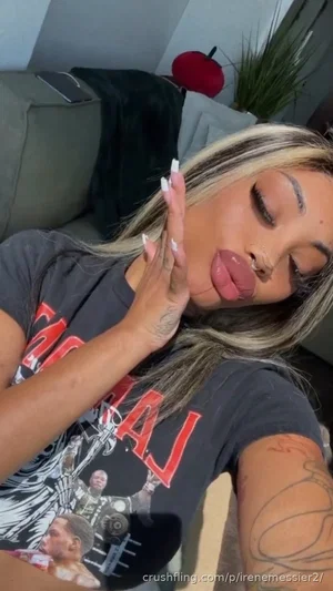 Woman with blonde highlights and tattoos wears graphic tee, posing indoors with hand near face.