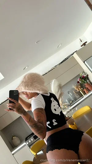Woman with tattoos in black and white crop top and lace underwear, taking mirror selfie in kitchen.