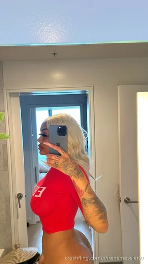 Woman with blonde hair and tattoos wears red crop top, taking mirror selfie in indoor setting.