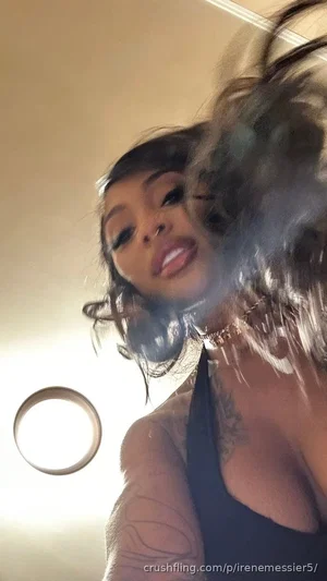 Woman with tattoos and dark hair in a black top, indoors, holding hair up.