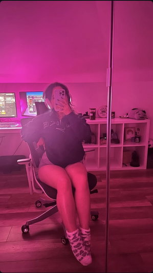 Woman in black hoodie and shorts sits in gaming chair, taking mirror selfie in pink-lit room.