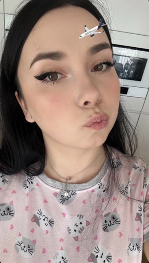 Woman with dark hair and makeup, wearing a pink cat-print shirt, making a playful face indoors.