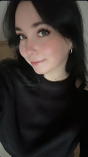 Woman with dark hair and green eyes wearing a black turtleneck, smiling softly indoors.