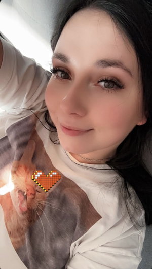 Woman with dark hair wearing white shirt with cat print, smiling in indoor selfie.