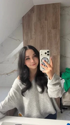 Woman with long dark hair in a gray sweater takes mirror selfie in bathroom.