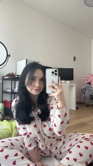 Woman with long dark hair in heart-patterned pajamas sitting indoors, taking mirror selfie.