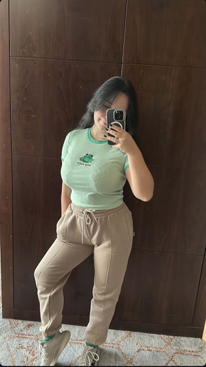 Woman in green t-shirt and beige pants poses for mirror selfie indoors.