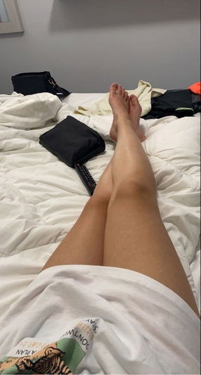 Woman in white shirt lying on bed, legs extended, bare feet visible, indoor setting.