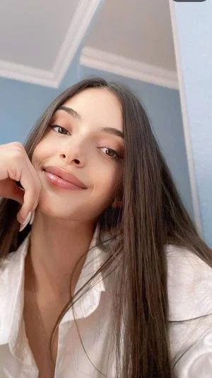 Woman with long dark hair in white shirt, smiling softly indoors.