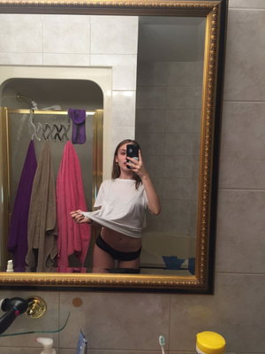 Woman in white shirt and black underwear taking mirror selfie in bathroom.