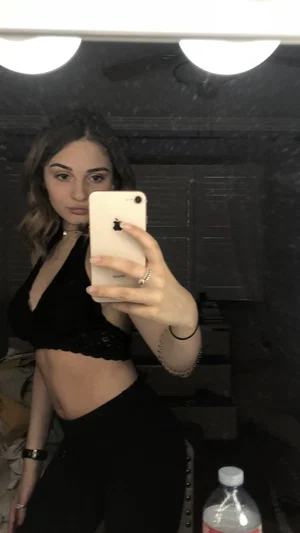 Woman in black crop top and pants takes mirror selfie in dimly lit room.
