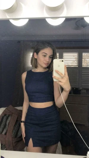 Woman in navy crop top and mini skirt takes mirror selfie in bedroom.