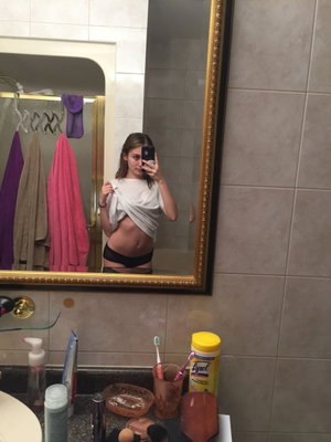 Woman in black underwear and white shirt partially lifted, taking mirror selfie in bathroom.