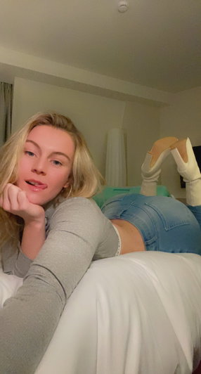 Blonde woman in gray top and jeans lying on bed, wearing white boots, indoor setting.