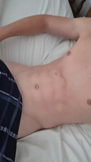 Man with bare torso lying on bed, wearing plaid shorts.