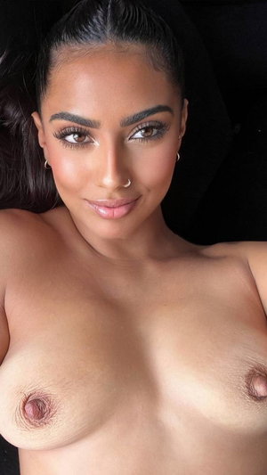 Curvy woman with dark hair and makeup, topless in dark setting.