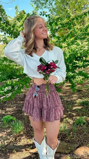 Woman in white crop sweater and floral skirt holding flowers outdoors in garden.