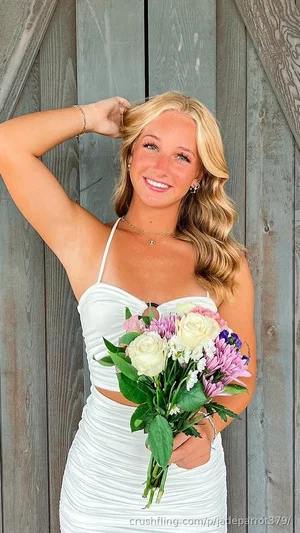 Blonde woman in white dress holding flowers, standing by wooden wall.