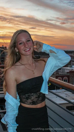 Blonde woman in black lace top and blue shirt, smiling on rooftop at sunset.