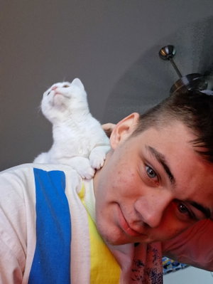 Man with short dark hair and a white cat on his shoulder, wearing a colorful striped shirt indoors.