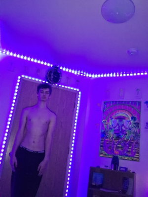 Shirtless man in black pants standing in a room with purple LED lights and a colorful poster.