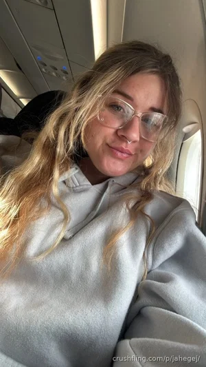 Woman with blonde hair and glasses wearing a gray hoodie on an airplane.