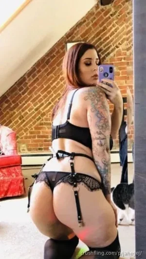Woman with tattoos in black lingerie and thigh-highs, taking mirror selfie in room with brick wall.