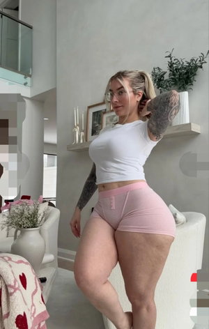 Woman in white crop top and pink shorts posing indoors, tattoos visible, modern living room setting.
