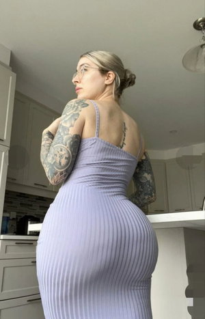 Woman with tattoos in a purple ribbed dress, standing in a kitchen.