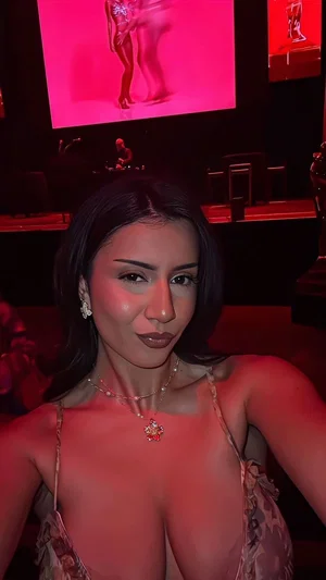 Woman with dark hair in a floral top, posing in a dimly lit room with red lighting.