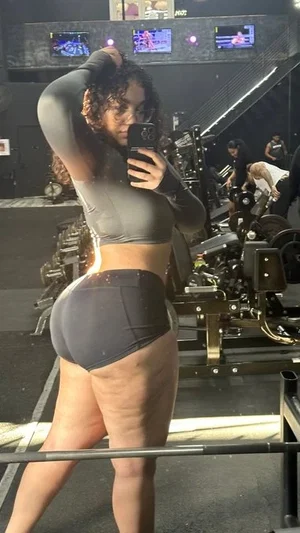Woman in gray crop top and black shorts taking mirror selfie in gym.