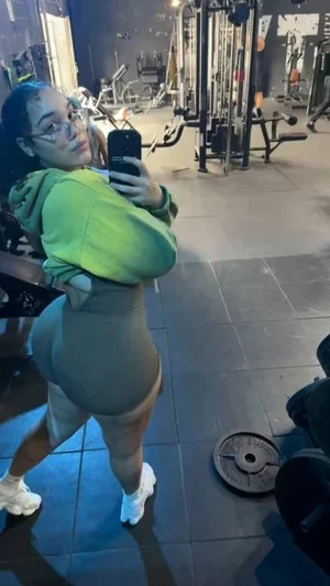 Woman in green hoodie and gray leggings takes mirror selfie in gym.