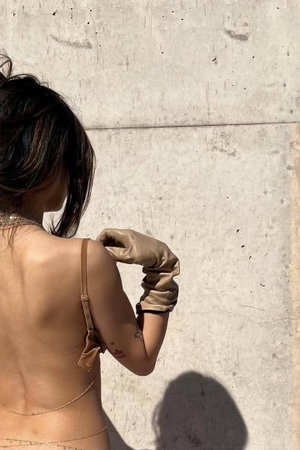 Woman in beige lingerie and gloves, back view, standing against concrete wall.