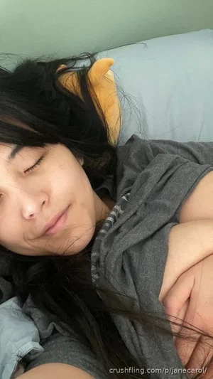 Woman with long dark hair sleeping in bed, wearing gray shirt, head on pillow.
