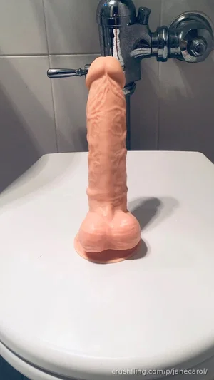 Pink flesh-toned dildo on white toilet lid near chrome faucet.