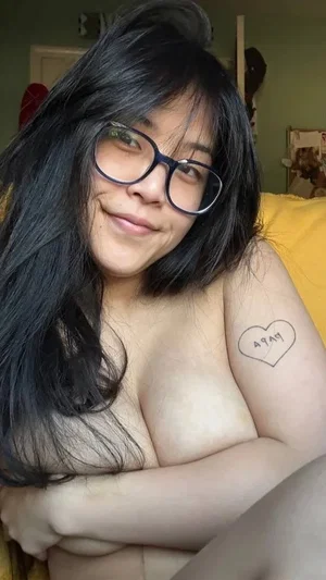 Woman with black hair and glasses, topless, covering chest with arms, heart tattoo on arm, indoors.