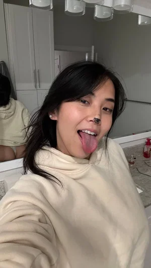 Woman with dark hair in a cream hoodie sticks tongue out in bathroom mirror selfie.