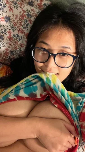 Woman with dark hair and glasses lying in bed, covering chest with colorful blanket.