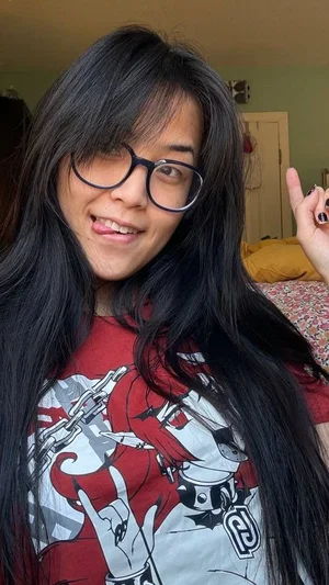 Woman with long black hair and glasses wearing a red graphic t-shirt, smiling in a bedroom.