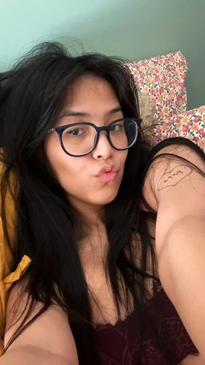 Woman with long black hair and glasses wearing a dark lace top, lying on a floral pillow indoors.