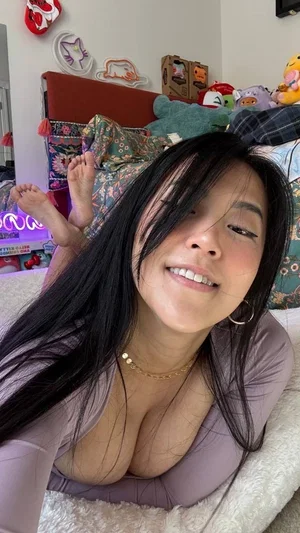 Woman with long black hair in a tight lavender top lying on a bed with plush toys and neon lights.