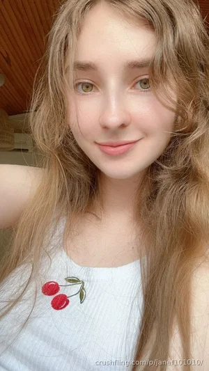 Woman with long blonde hair wearing white tank top with cherry design, indoor selfie.