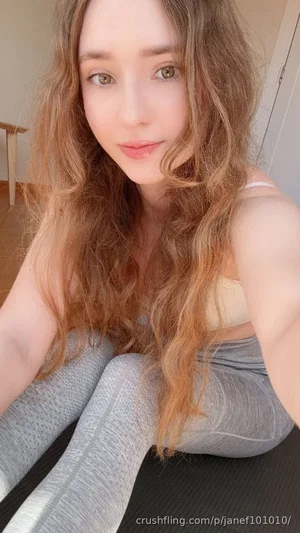 Woman with wavy brown hair in a beige top and gray leggings, sitting indoors on a mat.