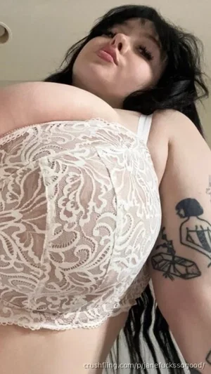Woman with dark hair and tattoos wears white lace bra, lying down indoors.
