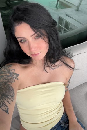 Woman with dark hair and tattoos wears yellow strapless top and jeans, sitting indoors.