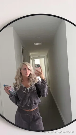 Blonde woman in gray hoodie and sweatpants takes mirror selfie in hallway.